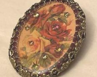 May include: An oval brooch featuring a central image of red and yellow roses. The image is surrounded by a dark red border and an ornate, textured gold-tone metal frame. Small, clear, sparkling rhinestones are embedded within the floral design.