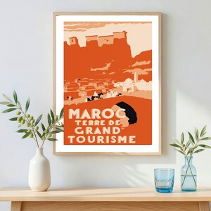 May include: A framed travel poster featuring a stylized orange and cream illustration of a Moroccan landscape with buildings and figures. The text MAROC TERRE DE GRAND TOURISME is printed in bold, light-colored capital letters at the bottom.