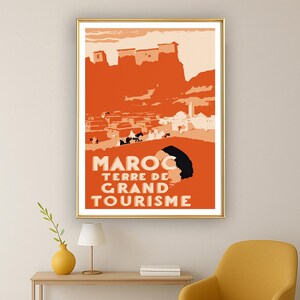 May include: A rectangular wall art print featuring a stylized orange and cream illustration of a Moroccan landscape with buildings and figures. The text MAROC TERRE DE GRAND TOURISME is printed in bold cream letters at the bottom of the poster.