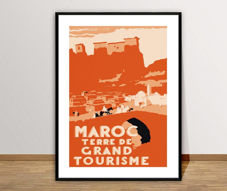 May include: A framed travel poster featuring a stylized orange and cream illustration of a Moroccan landscape with a fortress, a town, and figures on camels. The text MAROC TERRE DE GRAND TOURISME is printed in bold, light-colored capital letters.