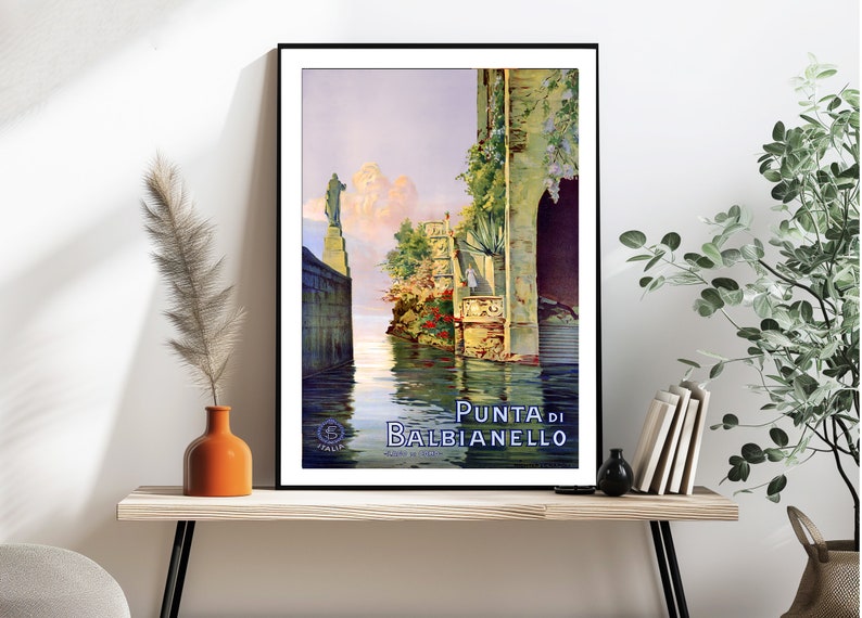 May include: Vintage travel poster featuring a scenic view of Punta di Balbianello, a beautiful Italian destination. The poster depicts a narrow waterway with a stone building on the right and a statue on the left. The water is calm and reflective, and the sky is a soft blue with fluffy clouds. The text "Punta di Balbianello" is prominently displayed in blue lettering.