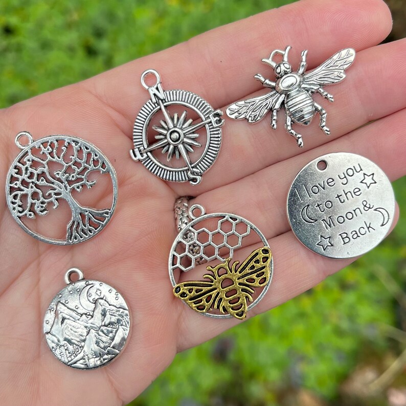 May include: A collection of silver and gold charm pendants featuring a compass, tree of life, honeycomb with a bee, a mountain scene, a bee, and a round charm with the text "I love you to the Moon & Back".