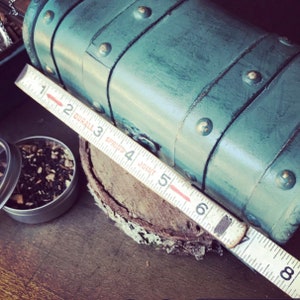 May include: A teal blue vintage-style wooden box with metal accents and a ruler measuring 6 inches. The box is sitting on a piece of tree bark.