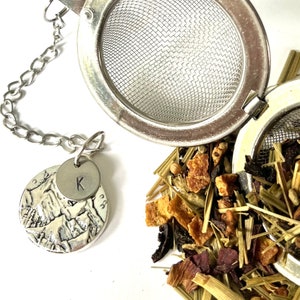 Pode incluir: Um infusor de chá de prata com uma corrente e um amuleto personalizado com a letra "K". O infusor está aberto e cheio de chá solto, incluindo frutas secas e ervas.