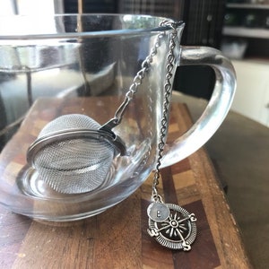 May include: A silver tea infuser with a mesh ball and a chain attached to a compass charm with the letter "E" on it. The infuser is hanging from the edge of a clear glass teacup.