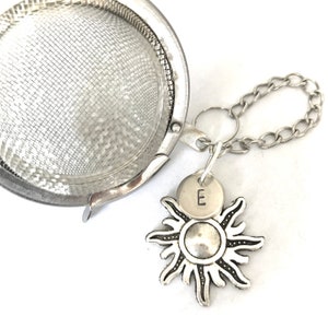 Pode incluir: Infusor de chá de prata com uma corrente e um amuleto de sol com a letra E em um disco pequeno.