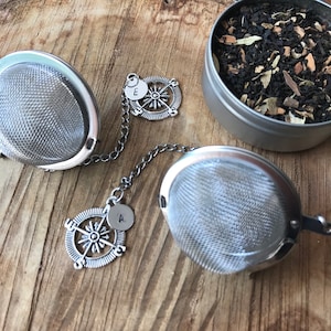 May include: Two silver tea infusers with a chain and compass charm. The charms have the letters 'A' and 'E' on them. The infusers are on a wooden surface.