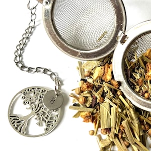 May include: A silver tea infuser with a chain and a tree of life charm with the letter B on it. The infuser is filled with loose leaf tea, including dried fruit, herbs, and spices.
