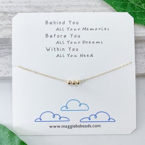 May include: A gold necklace with a delicate chain and four small gold beads. The necklace is displayed on a white card with the text "Behind You All Your Memories Before You All Your Dreams Within You All You Need" and the website address "www.maggiebsbeads.com".