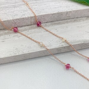 May include: A delicate rose gold chain necklace with alternating pink and white beads. The necklace is layered on a white wooden surface.