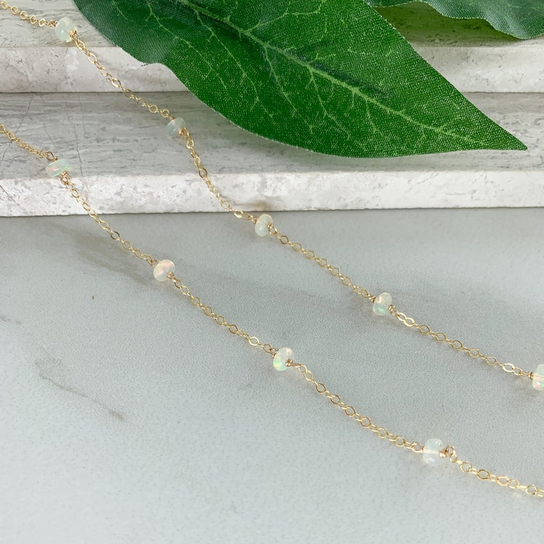 May include: A delicate gold chain necklace with small white opal beads. The necklace is on a white surface with a green leaf in the background.