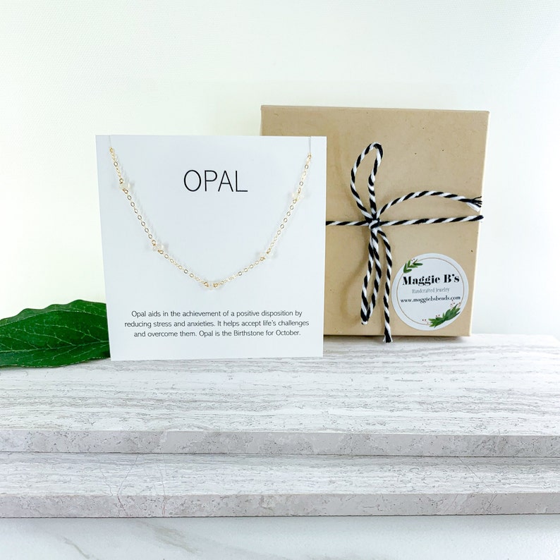 May include: A white card with the word "OPAL" printed in black. The card is sitting on a white wooden surface. A gold chain necklace with small white opal beads is displayed on the card. The necklace is packaged in a brown box with a black and white ribbon.
