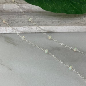 May include: A silver chain necklace with small white opal beads. The necklace is on a light gray surface.