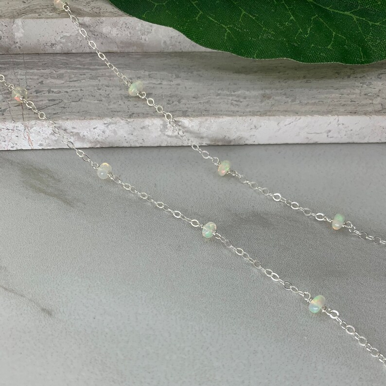 May include: A silver chain necklace with small white opal beads. The necklace is on a light gray surface.