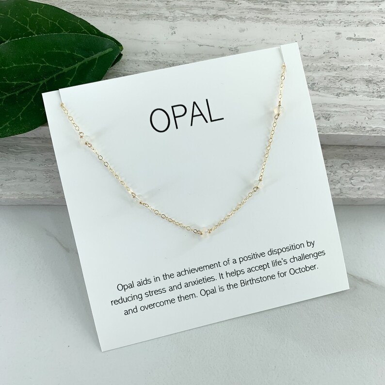 May include: A white card with the word "OPAL" printed in black. The card has a gold chain with small white opal beads around it. The card also has text about the meaning of opal and its birthstone status for October.