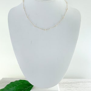 May include: A delicate gold chain necklace with small white beads. The necklace is displayed on a white mannequin bust.