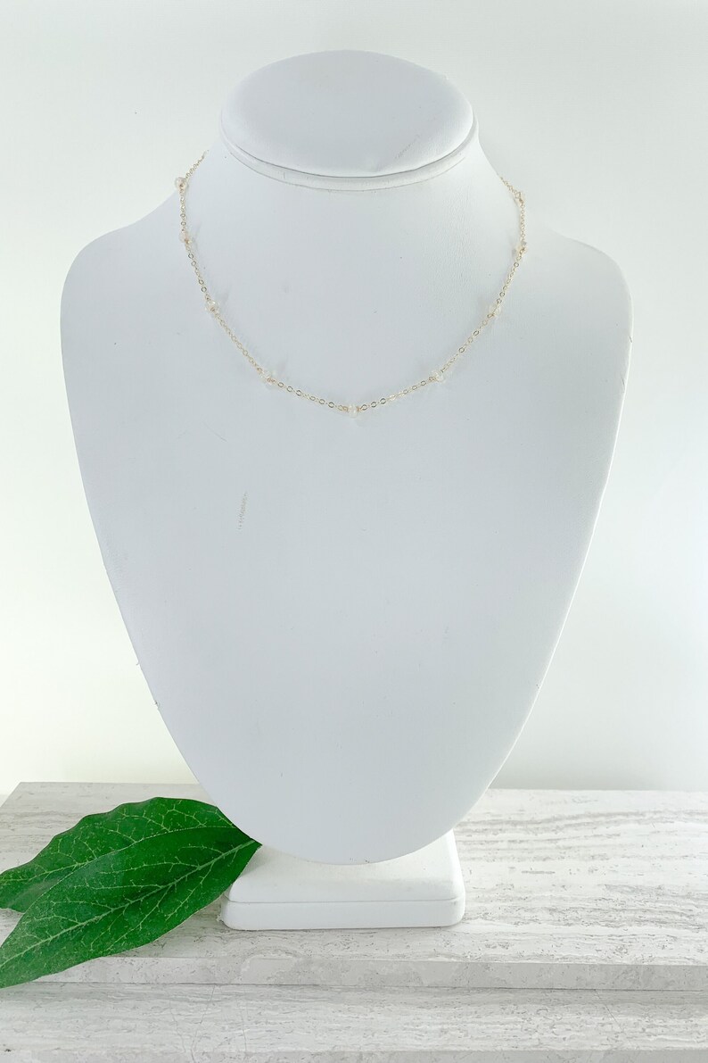 May include: A delicate gold chain necklace with small white beads. The necklace is displayed on a white mannequin bust.