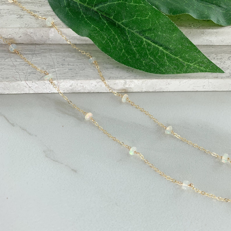 May include: A delicate gold chain necklace with small white opal beads. The necklace is displayed on a white surface with a green leaf in the background.