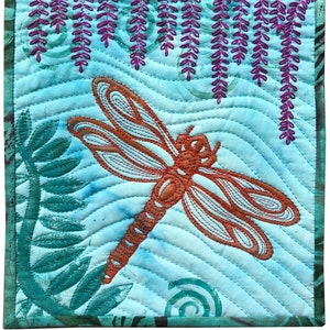May include: A quilted wall hanging with a blue background features a large orange dragonfly and purple wisteria flowers. The dragonfly is outlined in brown thread and the wisteria flowers are outlined in purple thread.