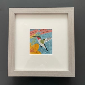 May include: A framed needlepoint embroidery of a hummingbird in flight with a yellow flower and pink and green foliage on a blue background.