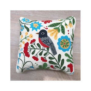 Puede incluir: Una almohada blanca con un diseño floral y un pájaro de colores. La almohada presenta un pájaro de pecho rojo con alas grises y un ojo negro, rodeado de hojas verdes, bayas rojas y flores azules y amarillas.