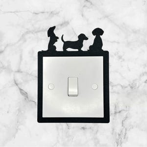 May include: A white light switch cover with a black border. Three dachshund silhouettes are on top of the cover. The dachshunds are facing different directions.