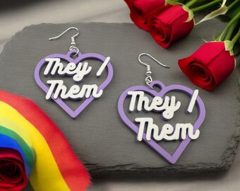 May include: A pair of purple heart-shaped earrings with the words "They/Them" in white lettering. The earrings are on a grey background with a rainbow ribbon and red roses.