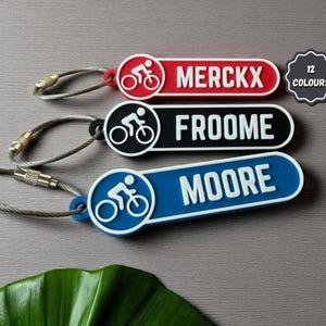 Könnte beinhalten: Drei Fahrrad-Schlüsselanhänger in Rot, Schwarz und Blau, jeweils mit einem Radfahrer-Symbol und einem Namen: MERCKX, FROOME und MOORE. Die Schlüsselanhänger haben eine silberne Drahtschlaufe zur Befestigung und den Text "12 COLOURS".