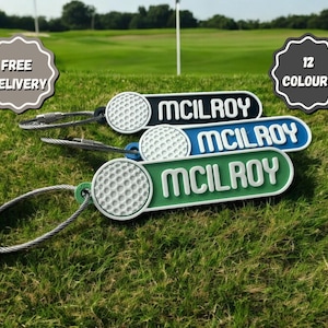 Peut inclure: Trois porte-clés en forme de balle de golf avec le nom "McIlroy" imprimé dessus. Les porte-clés sont en noir, bleu et vert. Les porte-clés sont sur une surface herbeuse verte. La livraison gratuite et 12 couleurs sont mentionnées sur l'image.