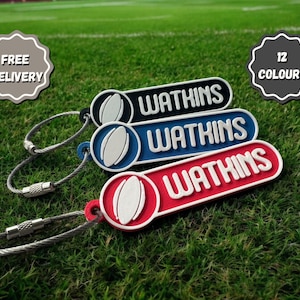 Op de afbeelding: Drie sleutelhangers met een rugbybalontwerp en het woord "Wathins" erop gedrukt. De sleutelhangers zijn zwart, blauw en rood. De sleutelhangers liggen op een groene grasmat.