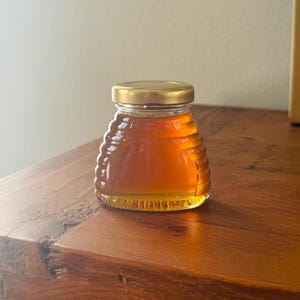 May include: A small glass jar with a gold lid filled with honey. The jar is shaped like a honeycomb and has a textured surface.