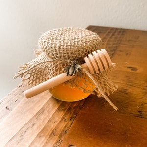 Puede incluir: Un pequeño tarro de vidrio de miel con una tapa de arpillera y un cucharón de miel de madera. El tarro tiene un pequeño amuleto de abeja de metal unido a la tapa.