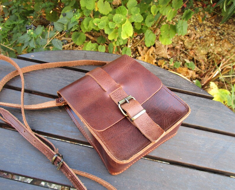May include: A brown leather crossbody bag with a flap closure and a metal buckle. The bag has a long adjustable strap.