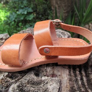 K&ouml;nnte beinhalten: Braune Ledersandalen mit Schnallenverschluss und gen&auml;hter Sohle. Die Sandalen befinden sich auf einer Holzoberfl&auml;che.