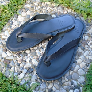 May include: Black leather flip-flops with a classic thong design. The sandals are shown on a stone surface with green grass. The flip-flops have a smooth, matte finish and are likely handmade. The size is 43.