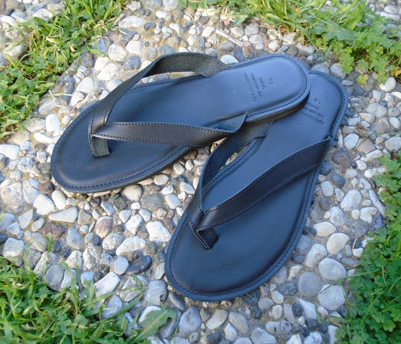 May include: Black leather flip-flops with a classic thong design. The sandals are shown on a stone surface with green grass. The flip-flops have a smooth, matte finish and are likely handmade. The size is 43.