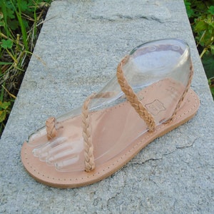 May include: Tan leather sandals with a braided strap design. The sandals have a thong toe strap and an ankle strap.