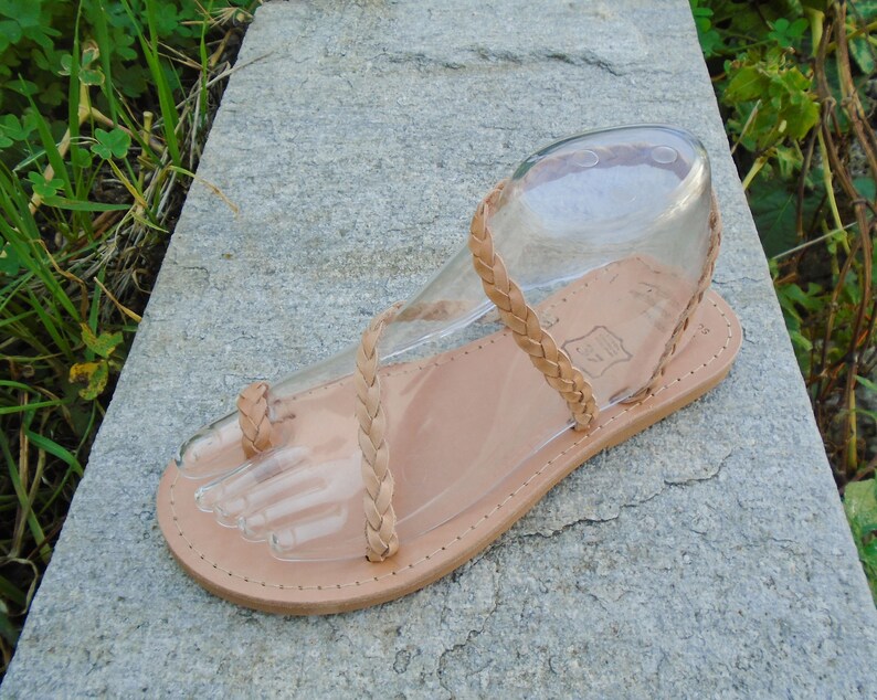 May include: Tan leather sandals with a braided strap design. The sandals have a thong toe strap and an ankle strap.