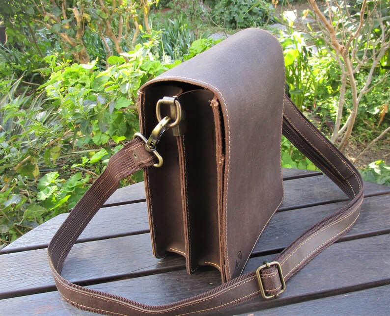 May include: A brown leather messenger bag with a long adjustable strap. The bag has a flap closure and a metal clasp.