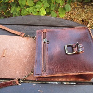 May include: A brown leather crossbody bag with a flap closure and a zippered pocket. The bag has a buckle on the front and a strap that can be adjusted.