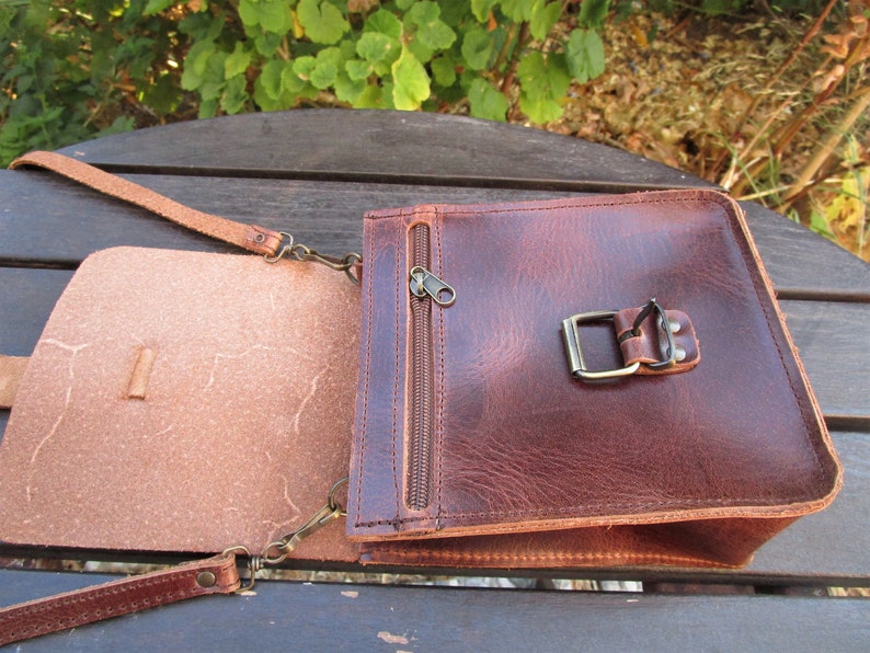 May include: A brown leather crossbody bag with a flap closure and a zippered pocket. The bag has a buckle on the front and a strap that can be adjusted.