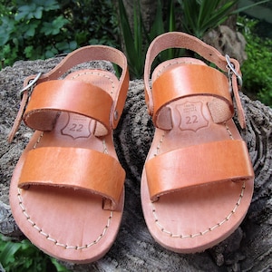 K&ouml;nnte beinhalten: Ein Paar braune Ledersandalen mit zwei Riemen und einem Schnallenverschluss. Die Sandalen haben einen gen&auml;hten Rand und ein Leder-Etikett mit dem Text "100% Echtleder 22" auf dem Riemen.