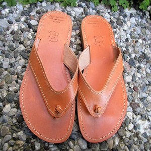 May include: A pair of brown leather thong sandals with a stitched design and the text "Apokoronos" on the top strap. The sandals are size 42.