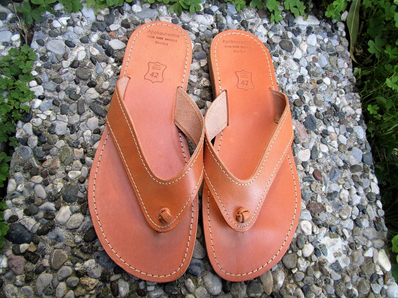 May include: A pair of brown leather thong sandals with a stitched design and the text "Apokoronos" on the top strap. The sandals are size 42.