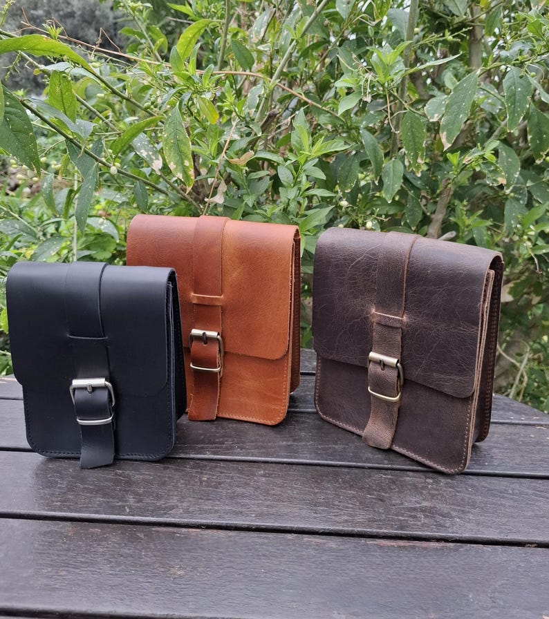 May include: Three leather bags in black, brown, and dark brown with a buckle closure. The bags are sitting on a wooden surface.