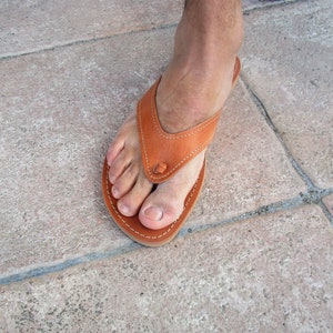 May include: A close-up of a brown leather thong sandal with a simple design. The sandal is worn on a foot with the toes visible.