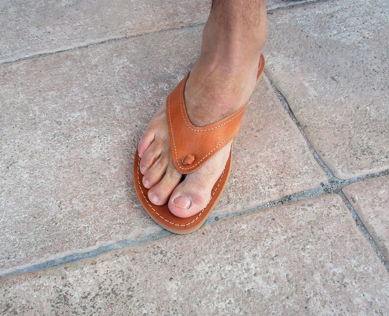 May include: A close-up of a brown leather thong sandal with a simple design. The sandal is worn on a foot with the toes visible.