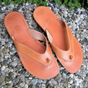 May include: A pair of brown leather sandals with a thong strap and a small knot on the strap. The sandals have a stitched edge and the text "Apokopaustos Hand Made Sandals Rhodes" is printed on the sole.