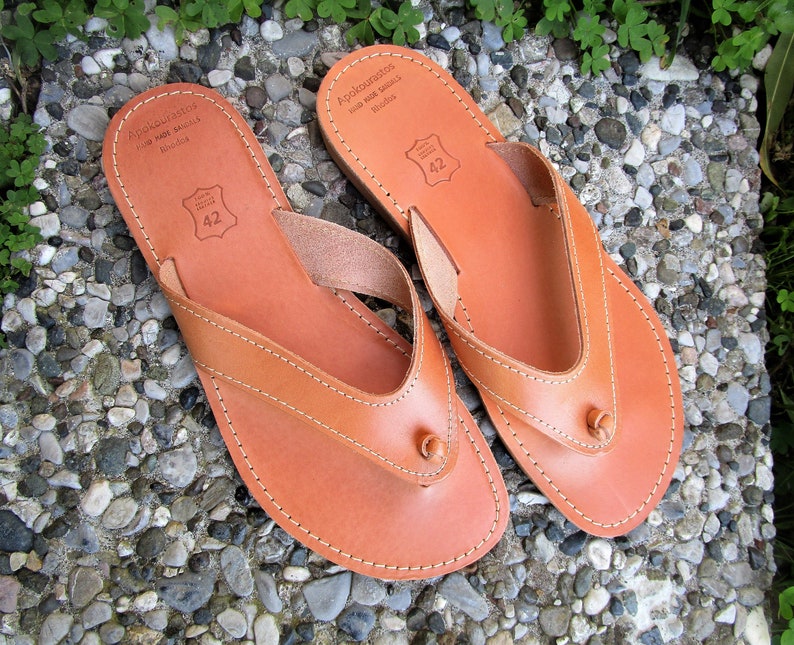 May include: A pair of brown leather sandals with a thong strap and a small knot on the strap. The sandals have a stitched edge and the text "Apokopaustos Hand Made Sandals Rhodes" is printed on the sole.