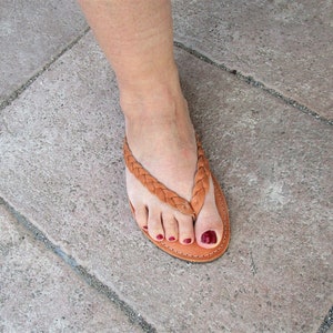 Pu&ograve; includere: Un paio di sandali in pelle marrone con un design a cinturino intrecciato. I sandali hanno una cinturino per le dita in stile thong e sono indossati su un piede con smalto rosso.